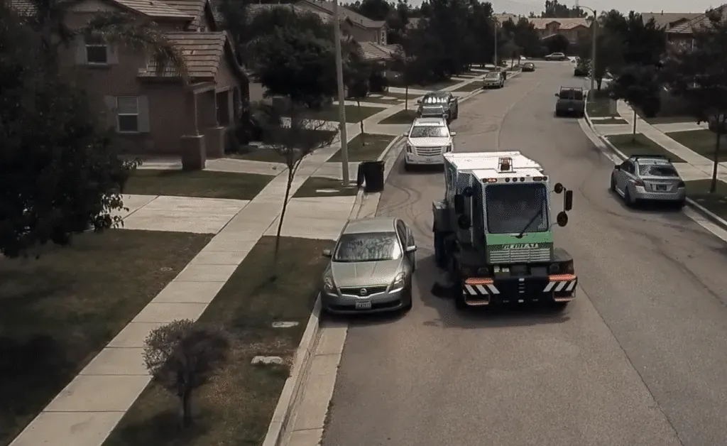 Street cleaning vehicle on residential street while parked cars line the curb.