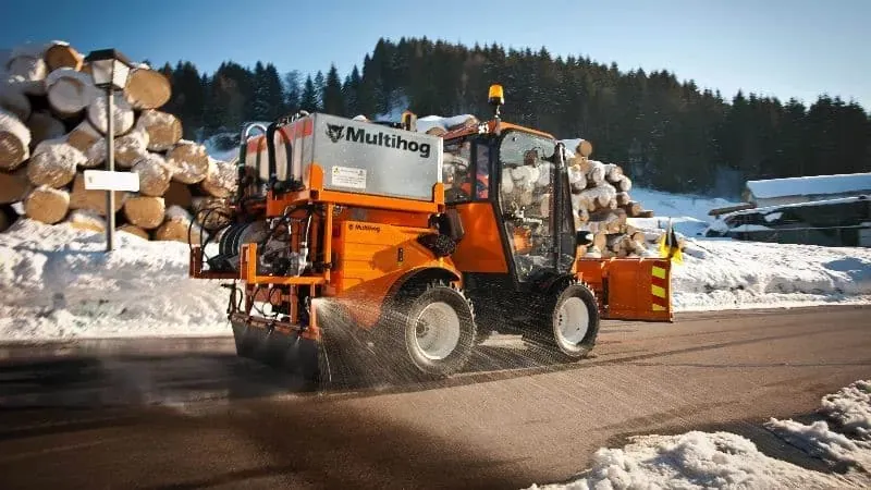 Multihog vehicle clearing snow while surrounded by logs in a winter landscape.